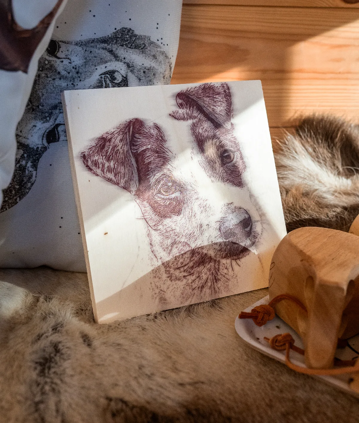 Träskiva med ett Jack Russell-porträtt i träbränningsstil, lutad mot en kudde med hundmotiv på en fäll i solljus, bredvid en träkåsa med läderband.